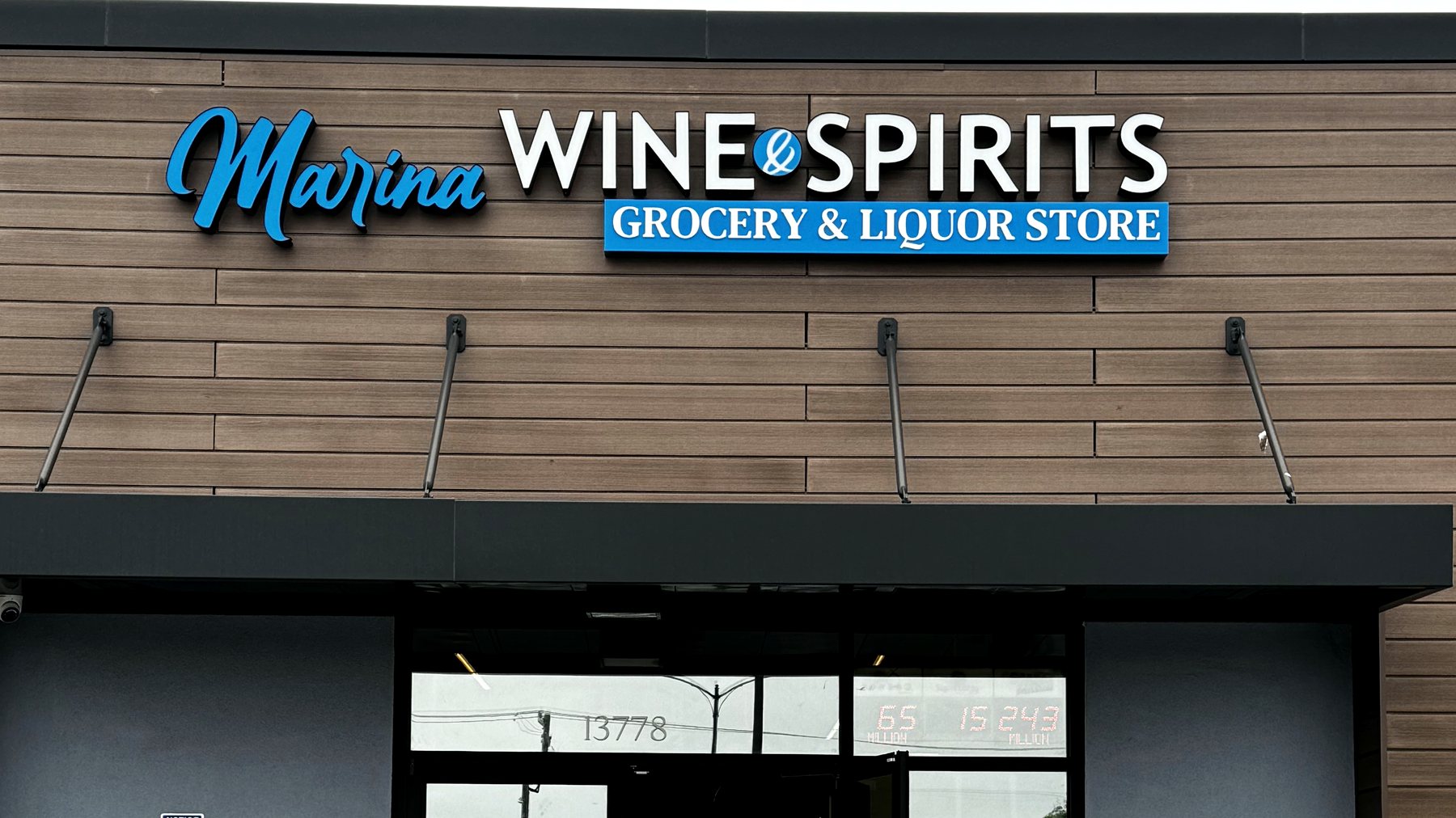 Close view of the Marina Wine & Spirits sign: blue script lettering reading Marina beside white capitals reading Wine and Spirits, over a blue banner reading Grocery and Liquor Store.